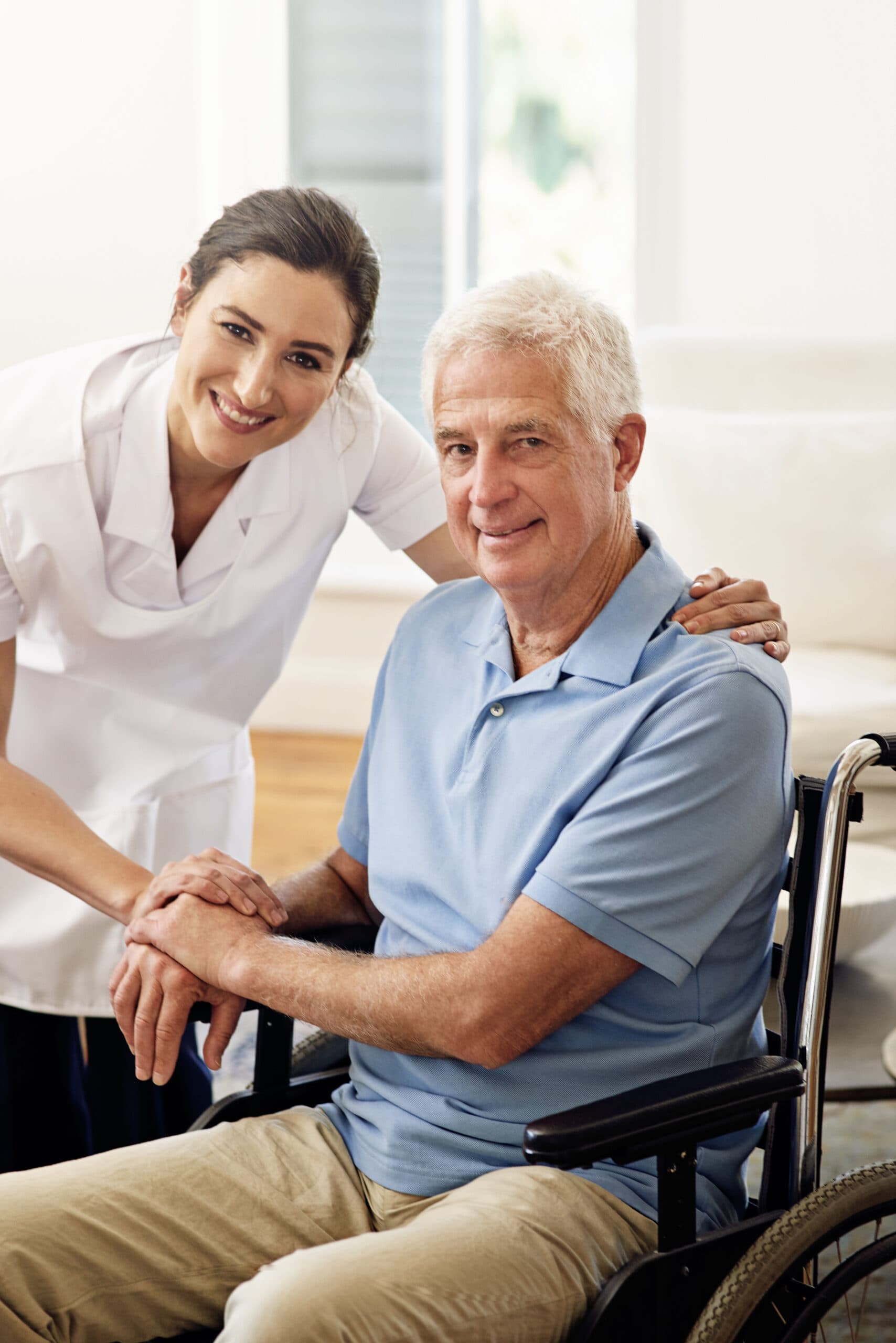 A smiling carer with her hands on the shoulders of an older man in a wheelchair